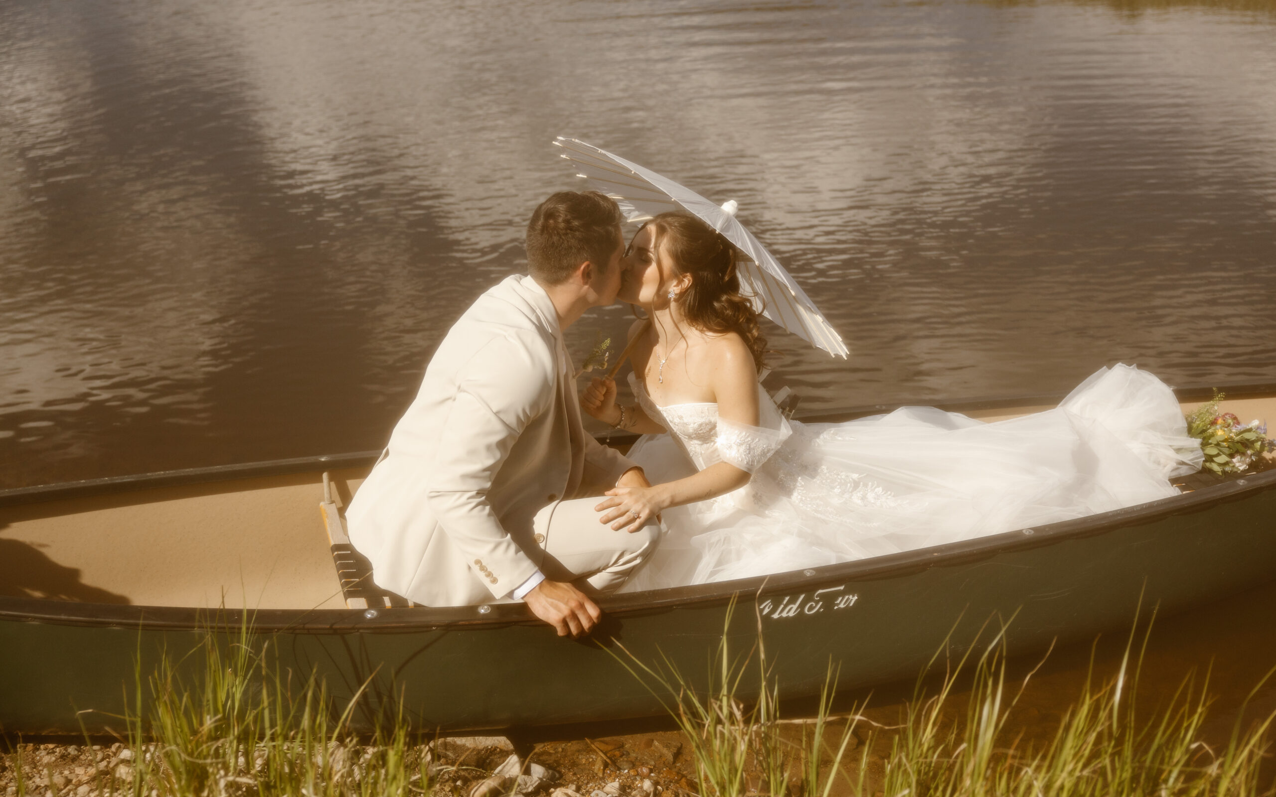 Colorado elopement couple kissing in a canoe on a calm lake, with the bride holding a white parasol and wearing an off-the-shoulder gown. A romantic and adventurous wedding moment on the water at Camp Hale in Colorado.