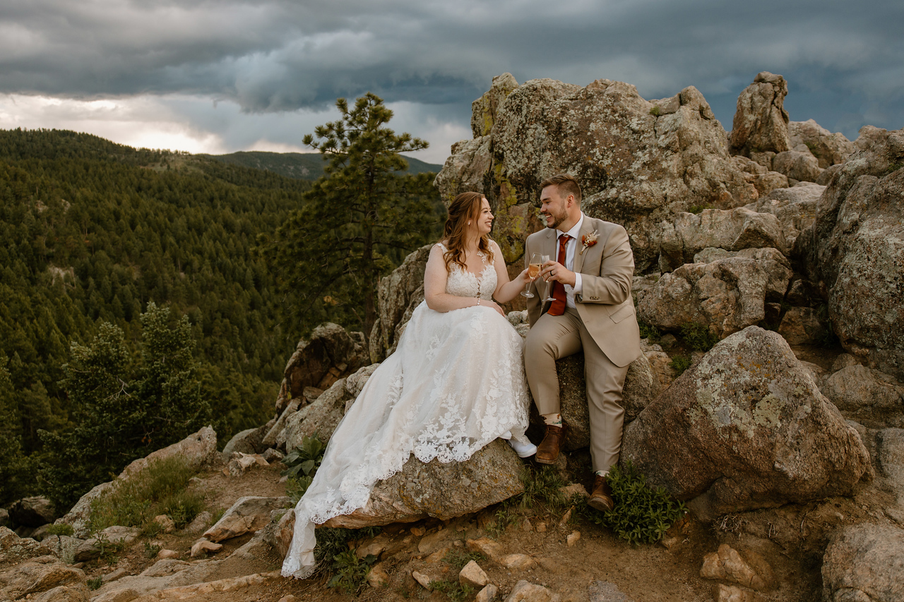 Bride and groom share a glass of champagne while sitting on a rock during their Colorado elopement celebration.