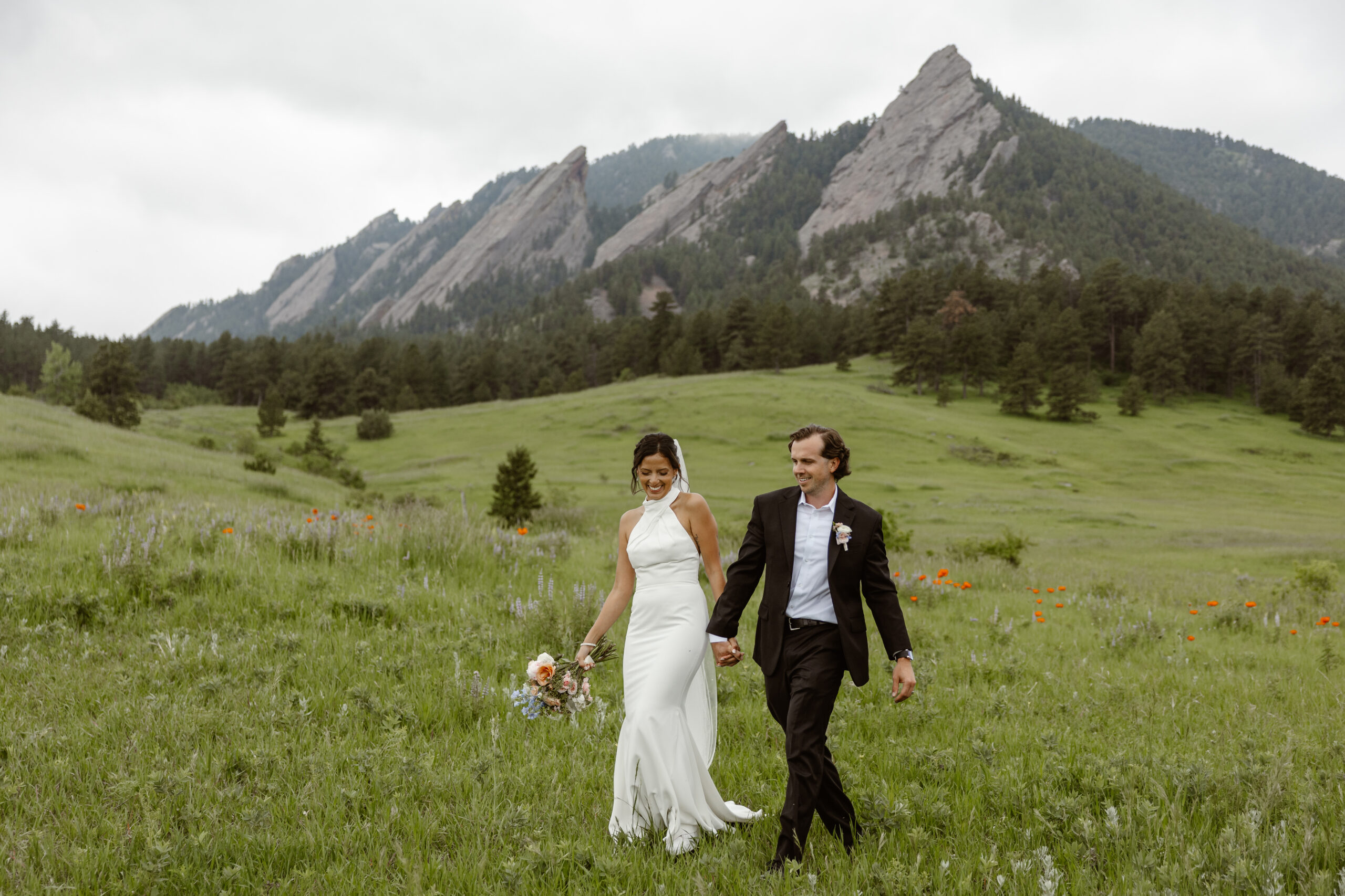 A bride and groom walking through a field of wildflowers with the Boulder flatirons in the background. The perfect location in Boulder for an intimate elopement!