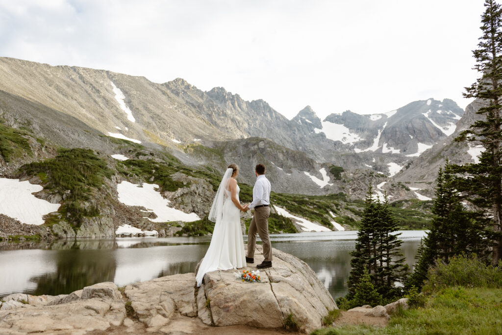 A bride and groom stand on a rock taking in the view of Indian Peaks Wilderness at Lake Isabelle in Colorado.