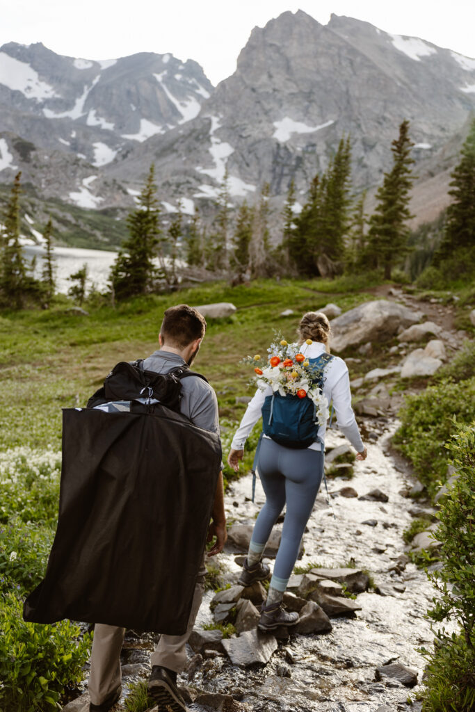 bride and groom hop over rocks to cross a river with their wedding attire strapped to their backpacks